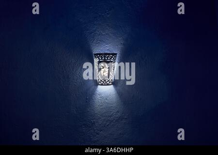 Traditional arabic style wall mounted lamp with light rays creating an abstract pattern on a dark blue textured wall background, Chefchaouen, Morocco Stock Photo