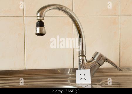 A sad face is drawn on paper standing near the kitchen sink in the kitchen in the apartment, no water Stock Photo