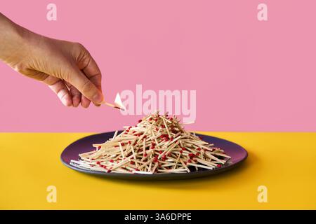 Hand holds burning matchstick above a large pile of matches on plate over vibrant background. Danger, risk concept Stock Photo