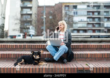 Design professional. Young freelance business woman, computer software developer, web designer, working remotely while sitting outdoor with her pet do Stock Photo