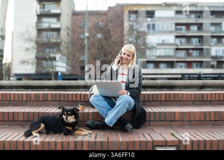 Design professional. Young freelance business woman, computer software developer, web designer, working remotely while sitting outdoor with her pet do Stock Photo