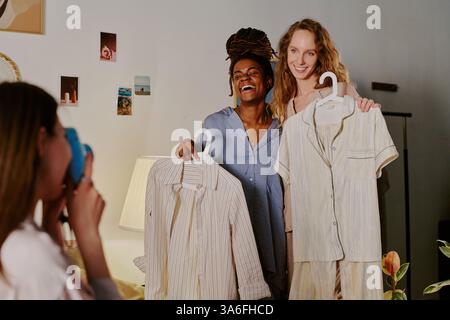 Low angle shot of two biracial best friends hugging each other and holding hangers with pajamas while posing for portrait together Stock Photo