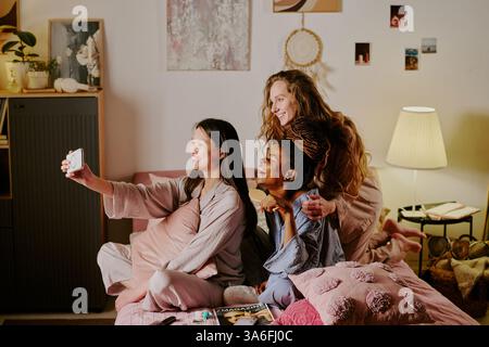 Wide shot of three beautiful biracial best friends wearing pastel colored pajamas taking selfies on double bed in girlish bedroom Stock Photo