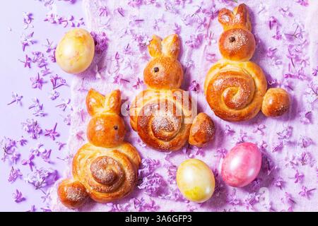 Easter composition made of sweet buns in the shape of a rabbit, eggs and lilacs. Yeast buns and painted eggs on a purple background, rustic style, sel Stock Photo