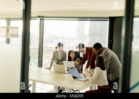 A diverse group of professionals engages in a dynamic brainstorming session around a conference table, sharing ideas while interacting with laptops an Stock Photo