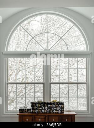 Wrapped presents on a pine chest in front of a transom window with a snow covered tree outside Stock Photo