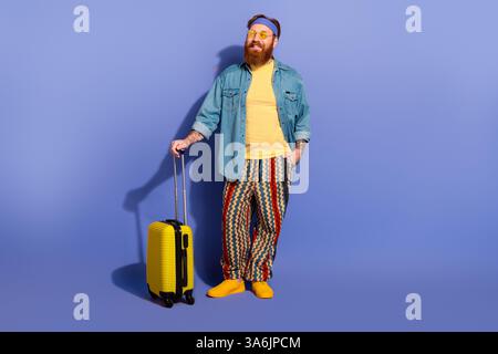Cheerful bearded man in vibrant casual attire with vintage accessories holding a yellow suitcase against a violet background Stock Photo