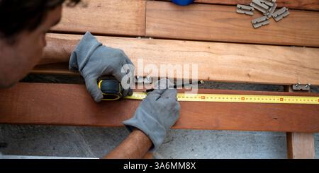 A craftsman wearing gloves uses a tape measure and pencil to mark a precise cut on a wooden deck, ensuring accuracy in construction. Stock Photo