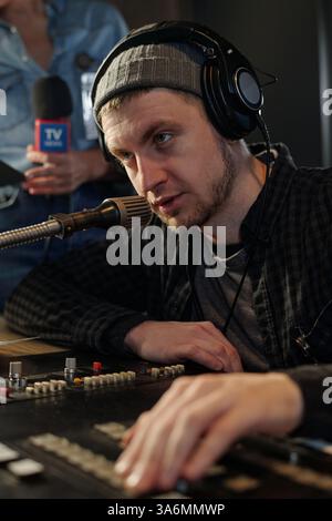 Young man in headphones speaking in microphone and mixing sounds on console while checking or adjusting equipment Stock Photo