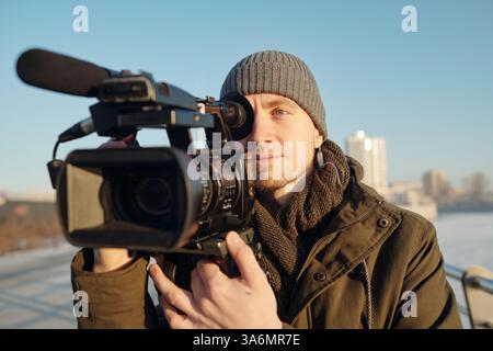 Male operator or videographer shooting new footage or reportage on video camera while standing in urban environment Stock Photo
