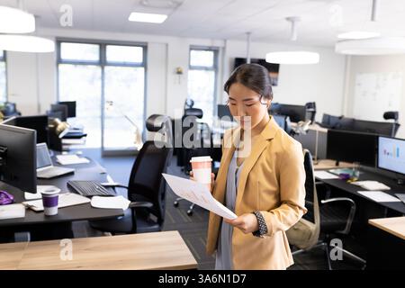 Businesswoman reviewing documents while holding coffee cup in modern office, copy space Stock Photo