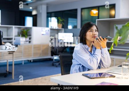 Asian woman in office using smartphone for voice message, looking thoughtful, copy space Stock Photo