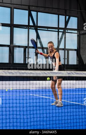 Young woman playing padel tennis on indoor court, focusing on hitting ball Stock Photo