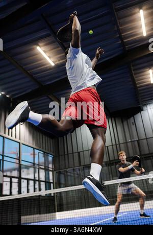 African American man jumping to hit ball during intense padel tennis match indoors Stock Photo