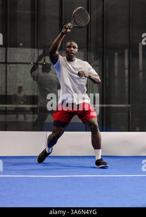 African American man playing padel tennis, focusing on hitting ball indoors Stock Photo