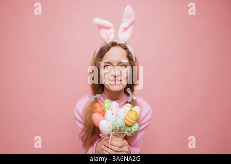 Happy woman in bunny ears holding a bouquet of colorful Easter eggs, smiling playfully. Festive spring mood, holiday joy, and seasonal celebration Stock Photo