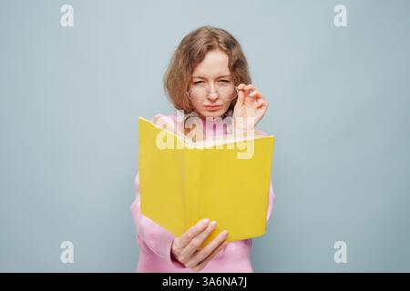 Serious woman with poor eyesight in glasses reading yellow book, adjusting glasses, concentrated, wearing pink sweater, over pastel blue background. Stock Photo