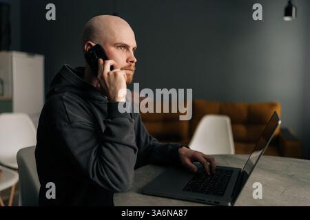 Side view of confident bearded bald freelancer wearing dark hoodie working remotely talking smartphone while typing on laptop computer in minimalist Stock Photo