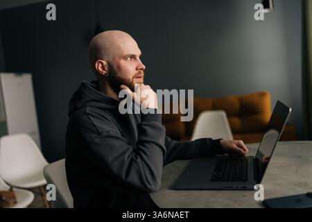 Thoughtful freelancer remote working on laptop computer sitting at table in home office, resting hand on chin, deep in thought looking away. Stock Photo