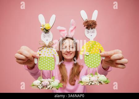 Smiling woman in pink holds handmade Easter bunny decorations, wearing bunny ears headband, against pastel pink background. Stock Photo