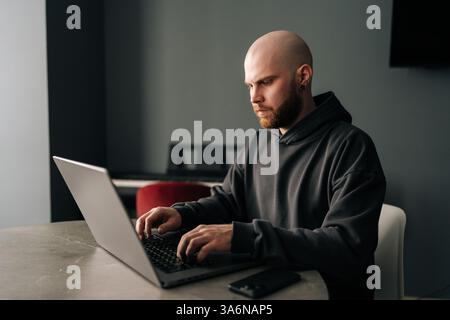 Side view of focused male programmer with beard and hoodie diligently typing on laptop computer sitting at table at modern home office, developing Stock Photo