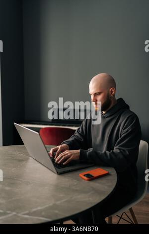 Vertical portrait of young bald programmer with beard and ear piercing sitting at round table in home office, concentrating on work on laptop looking Stock Photo