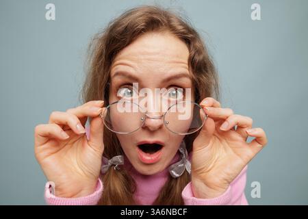 Surprised young woman lifting round glasses in shock, eyes wide open, wearing pink sweater, excited expression, amazed reaction, studio shot. Stock Photo