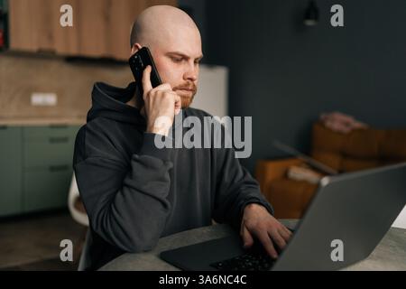 Serious freelancer male working from remote home office, talking on mobile phone and using laptop computer, managing business and multitasking in home Stock Photo