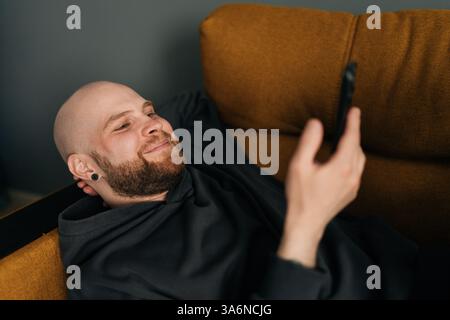 Close-up face of cheerful man with beard wearing dark hoodie lying comfortably on sofa using mobile phone, enjoying leisure time at home. Stock Photo