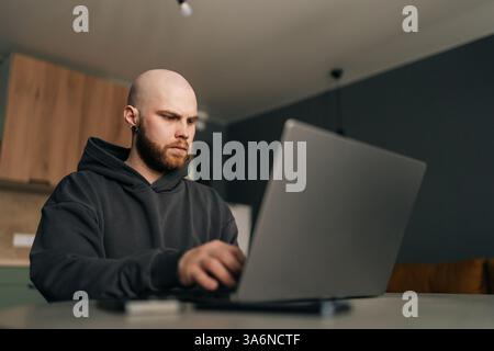 Low-angle view of focused programmer wearing dark hoodie typing code on laptop computer, sitting at table near bright window in contemporary urban Stock Photo