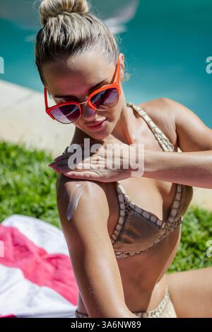 Woman in sunglasses applying sunscreen by pool, enjoying sunny day outdoors Stock Photo
