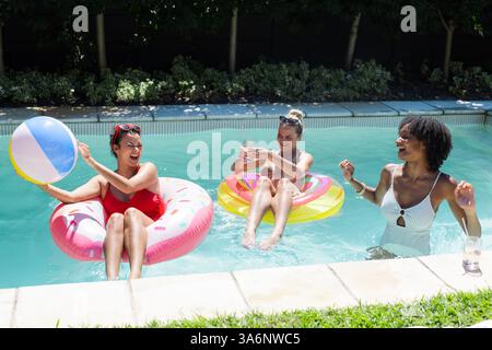 Diverse female friends enjoying pool party, laughing and playing with beach ball on floats Stock Photo