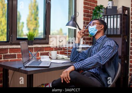 African american manager seated in brick wall office looking pensive and tapping his forehead with a pen. Businessman with face mask, gazing outside the window and thinking deeply on company projects. Stock Photo