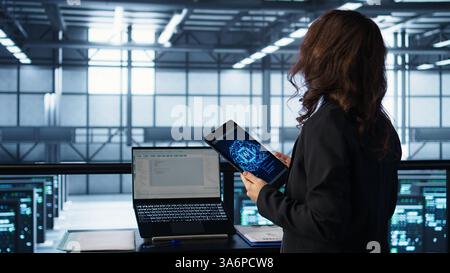 Technician in data center with tablet integrating AI driven automation tools, running predictive maintenance routines. Woman ensuring continuous uptime of server room, managing data flows, camera B Stock Photo