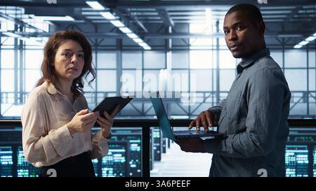 Data scientists in server room using laptop and tablet, executing security patches, verifying firewall configurations. People performing equipment vulnerability scans in data center, camera B Stock Photo