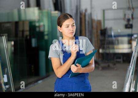 Female technician inspecting glass panels and writing notes in workshop Stock Photo