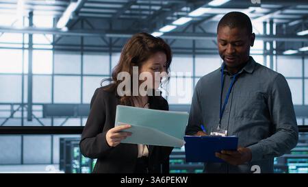 IT professionals in data center reviewing reports and files, crosschecking network performance metrics and security logs. Server room workers doing troubleshooting to prevent downtime, camera A Stock Photo