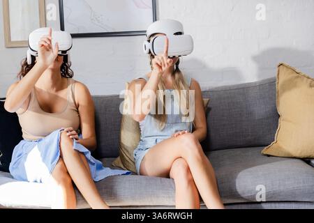 Women using VR headsets on sofa, exploring virtual reality together Stock Photo