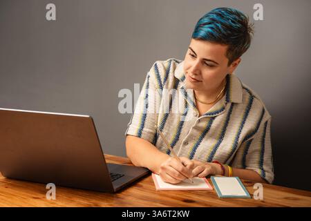 Smiling young woman with short blue hair taking notes while attending a video call on her laptop at a wooden desk. Remote work and digital planning co Stock Photo
