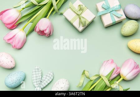 Easter frame made of colorful eggs, pink tulips and gift boxes on light green background with copy space in the center Stock Photo
