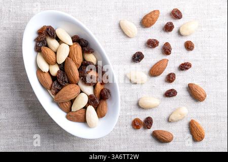 Almonds and raisins, a trail mix in an oval white bowl, on linen fabric. Also known as student food since 17th century. Stock Photo
