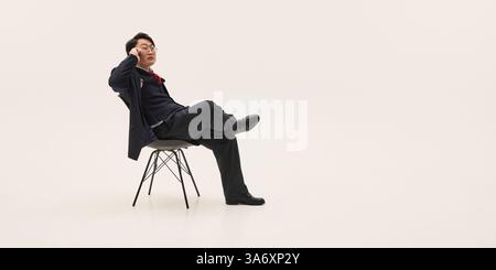 Young Asian man dressed stylish formal attire sitting on chair and talking on phone with serious expression against white background. Stock Photo