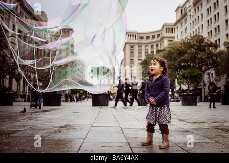 Toddler girl playing outdoors with soap bubbles in a public square in the european city of Barcelona. Children playing outside Stock Photo