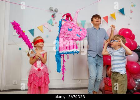 Child in a blindfold and dress aiming stick at unicorn pinata Stock Photo