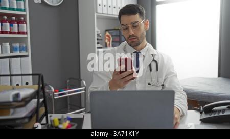 Young hispanic man wearing lab coat using smartphone at clinic desk, surrounded by medical equipment and files, in a modern hospital room setting with Stock Photo