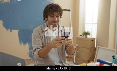 Young man smiling while using smartphone in a newly painted living room surrounded by moving boxes and paint supplies, suggesting a fresh start or new Stock Photo