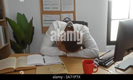 Young man exhausted in an office setting, resting head on desk next to open books, computer and red coffee mug Stock Photo