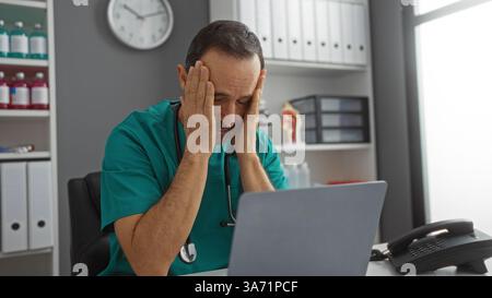 Hispanic male doctor in hospital room experiencing stress while sitting at a desk with hands on head, surrounded by office supplies and medical equipm Stock Photo