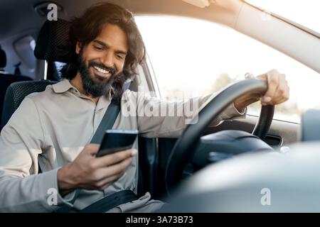 Unsafe driving. Cheerful indian man using smartphone while driving car Stock Photo