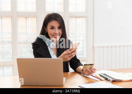 Young asian woman holding credit card and using smartphone for purchase product on internet in home office. Online shopping concept. Stock Photo